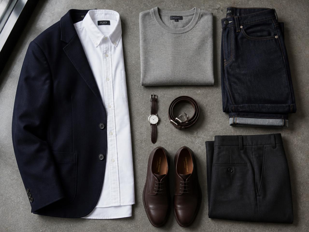 Flat lay of a men's capsule wardrobe: navy blazer, white oxford shirt, grey knit, dark denim, charcoal trousers, brown derbies, belt and watch